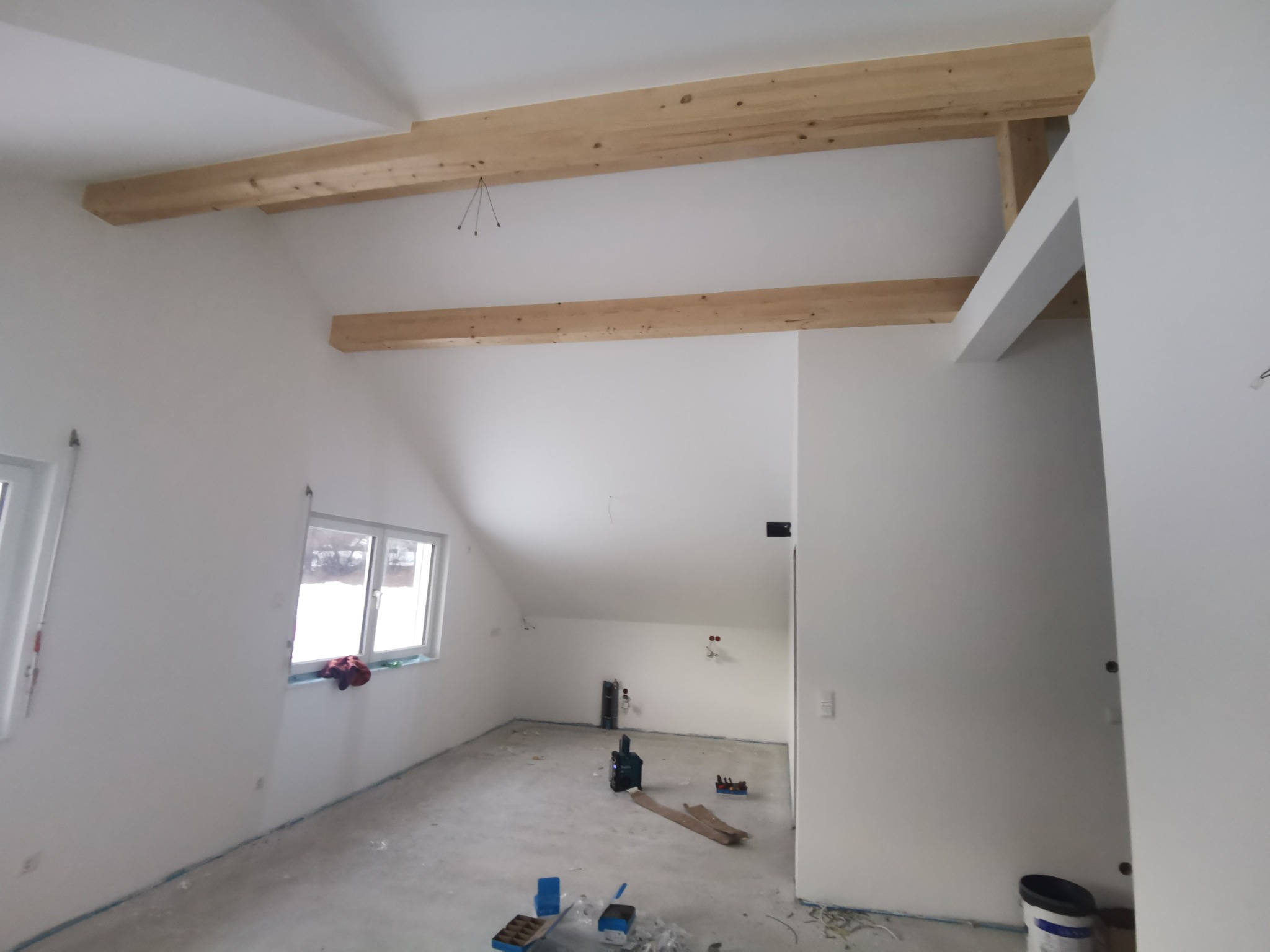 Poddasze po remoncie: białe ściany, drewniane belki stropowe, okno. Widoczne narzędzia i materiały budowlane. Minimalistyczny, surowy styl wykończenia.