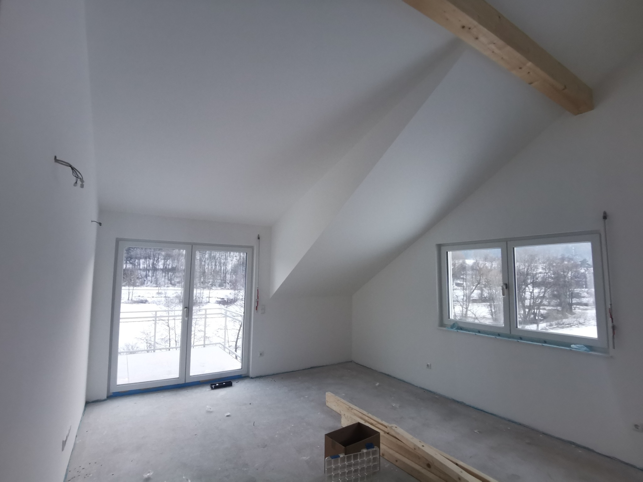 Poddasze po gipsowaniu ścian i sufitu, z oknami wychodzącymi na zimowy krajobraz. Widoczne drewniane belki i przygotowane materiały budowlane na podłodze.