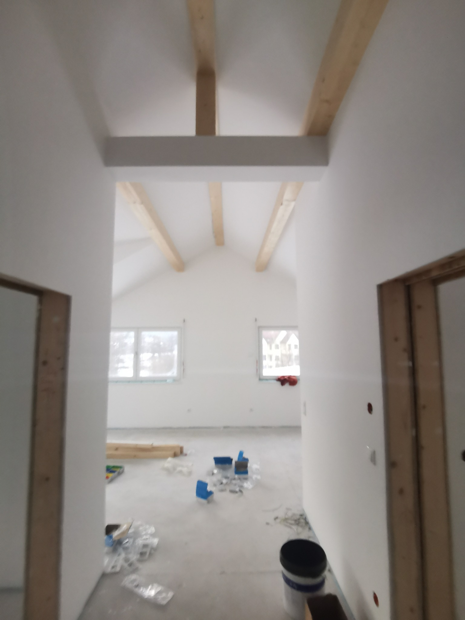Wnętrze w trakcie prac wykończeniowych: białe ściany, drewniane belki stropowe, okna. Widoczne narzędzia i materiały budowlane na podłodze. Surowy stan, przygotowanie do dalszych prac.