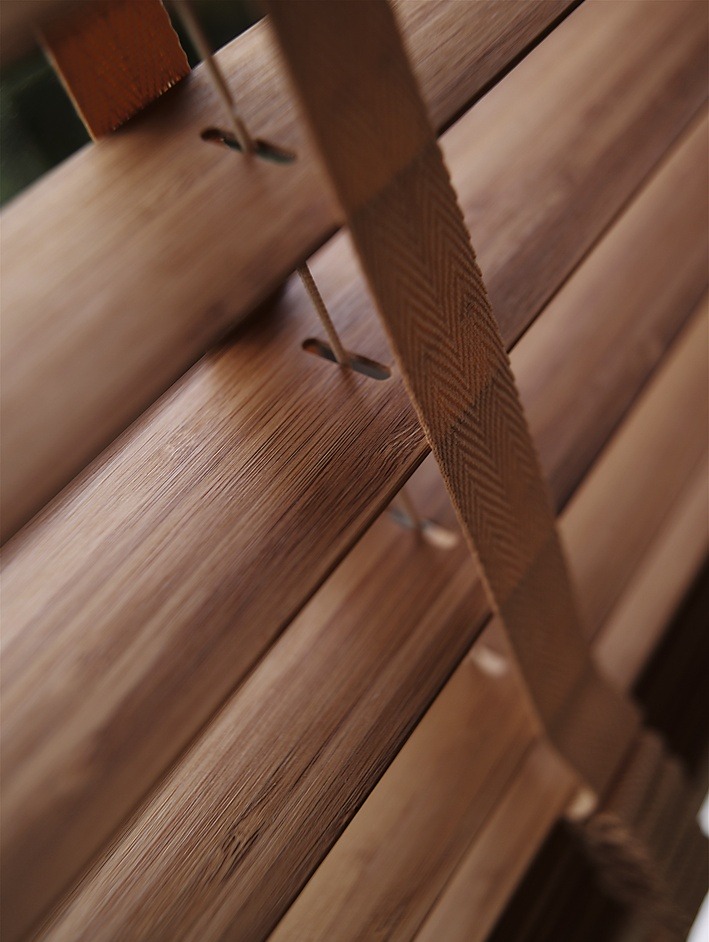 Fragment drewnianych żaluzji z bliska, widoczne poziome lamele połączone taśmą z wzorem w jodełkę i sznurkami.