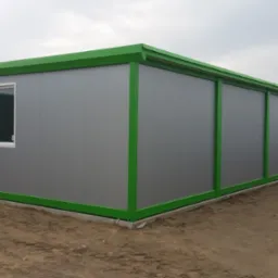 Długi, szary pawilon biurowy z zielonymi ramami okiennymi i obramowaniem dachu, ustawiony na placu budowy z widocznym piaskiem.