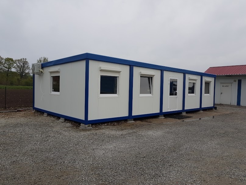 Prostopadłościenny moduł biurowy z klimatyzacją, białymi ścianami i niebieskimi obramowaniami, ustawiony na żwirowym podłożu, z widocznym budynkiem biurowym w tle.