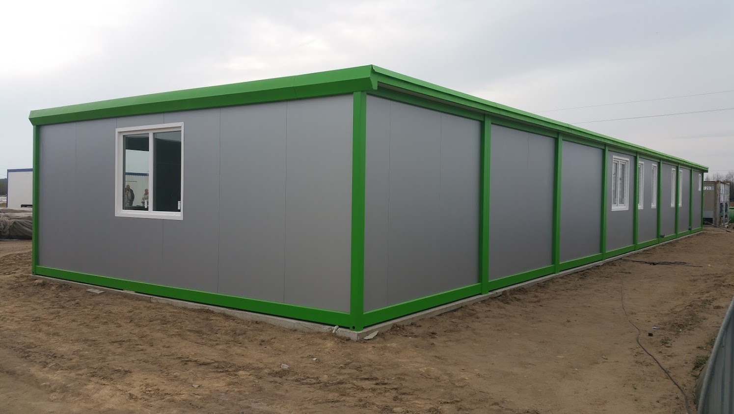 Długi, szary pawilon biurowy z zielonymi ramami okiennymi i obramowaniem dachu, ustawiony na placu budowy z widocznym piaskiem.