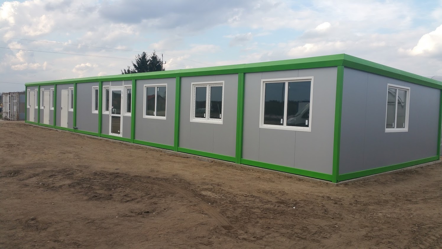 Długi, szary budynek modułowy z zielonymi obramowaniami okien i drzwi, widoczny na placu budowy z ziemistym podłożem i fragmentem kontenera w tle.