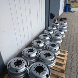 Rząd srebrnych felg do ciężarówek, świeżo po obróbce, ułożonych na drewnianych paletach przed budynkiem z szarej blachy falistej.