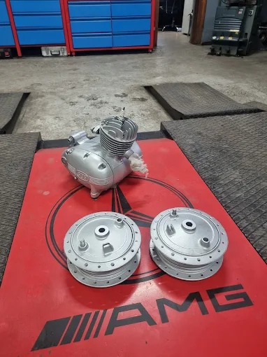 Silnik motocyklowy i dwie piasty po renowacji, pomalowane na srebrno, leżące na czerwonym stole warsztatowym z logo AMG.