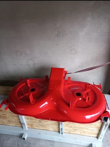 Czerwona obudowa kosiarki po renowacji, leżąca na drewnianej palecie, widok z boku na tle szarej ściany.