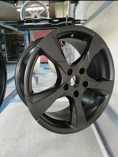 Matowa, grafitowa felga aluminiowa zawieszona na metalowym stojaku po aplikacji nowej powłoki. Widoczne detale mocowania i logo producenta felgi.