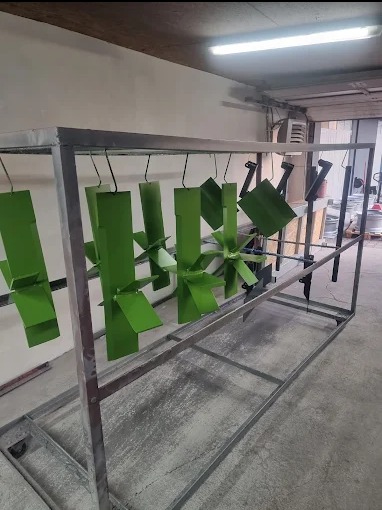 Elementy metalowe, niektóre w kolorze zielonym, inne czarne, zawieszone na metalowej konstrukcji w hali produkcyjnej, prawdopodobnie po procesie nakładania farby.