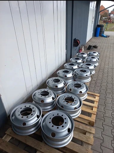 Rząd srebrnych felg do ciężarówek, świeżo po obróbce, ułożonych na drewnianych paletach przed budynkiem z szarej blachy falistej.