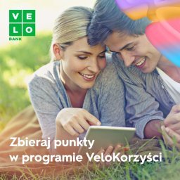 VeloBank S.A. - Uśmiechnięta para leżąca na kocu na trawie, korzystająca z tabletu, logo VeloBank w lewym górnym rogu, tekst 'Zbieraj punkty w programie VeloKorzyści'.