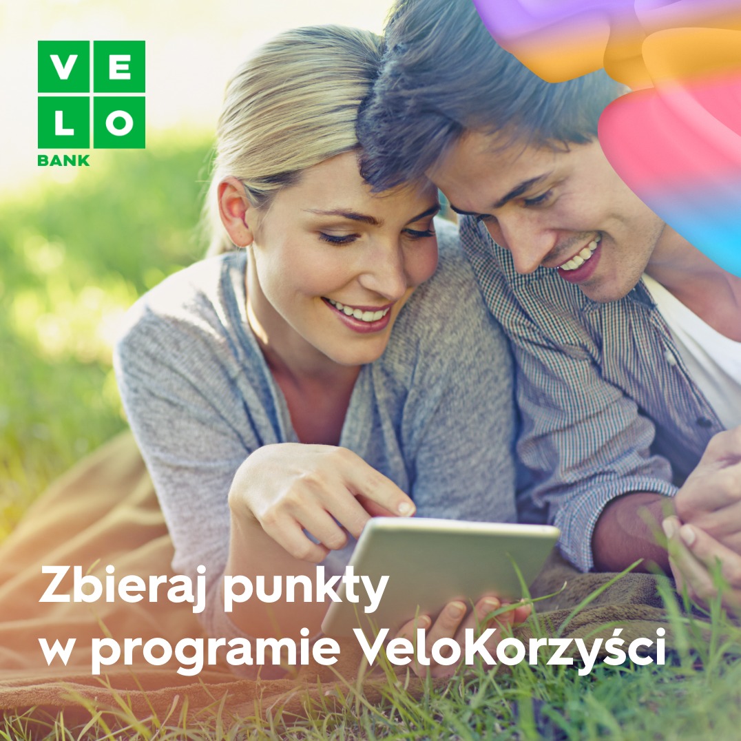 Uśmiechnięta para leżąca na kocu na trawie, korzystająca z tabletu, logo VeloBank w lewym górnym rogu, tekst 'Zbieraj punkty w programie VeloKorzyści'.