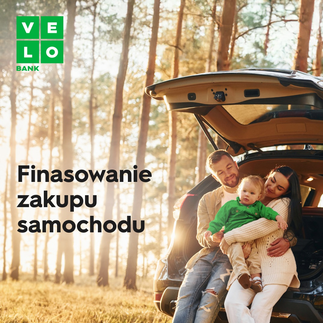 Rodzina z małym dzieckiem siedzi przy otwartym bagażniku czarnego SUV-a na tle leśnego krajobrazu w słoneczny dzień. Logo VeloBank w lewym górnym rogu.
