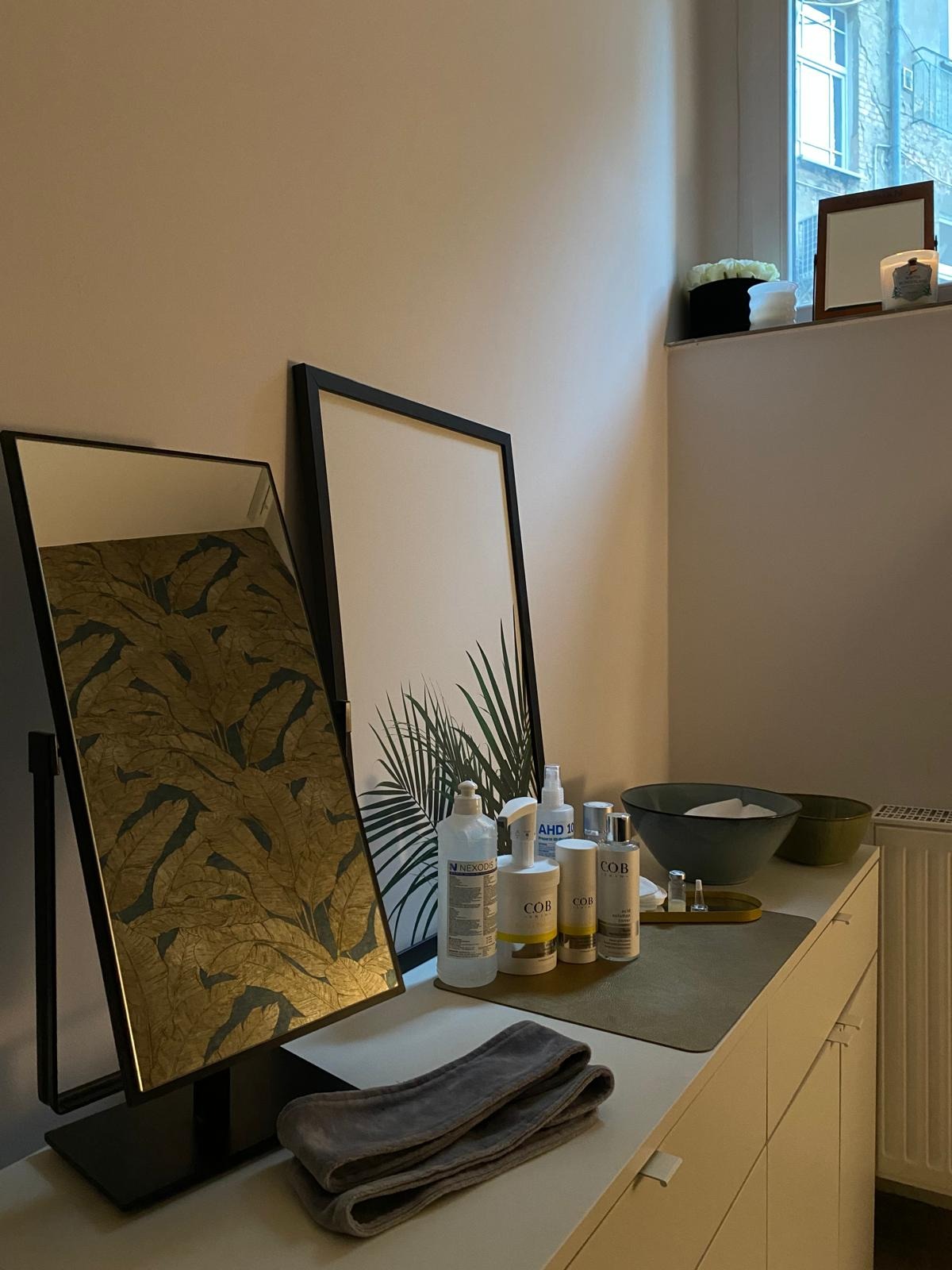 Przygotowane stanowisko do zabiegu: lustro z motywem liści, grafika z palmą, kosmetyki, miseczki i ręcznik na białej komodzie, w tle okno.