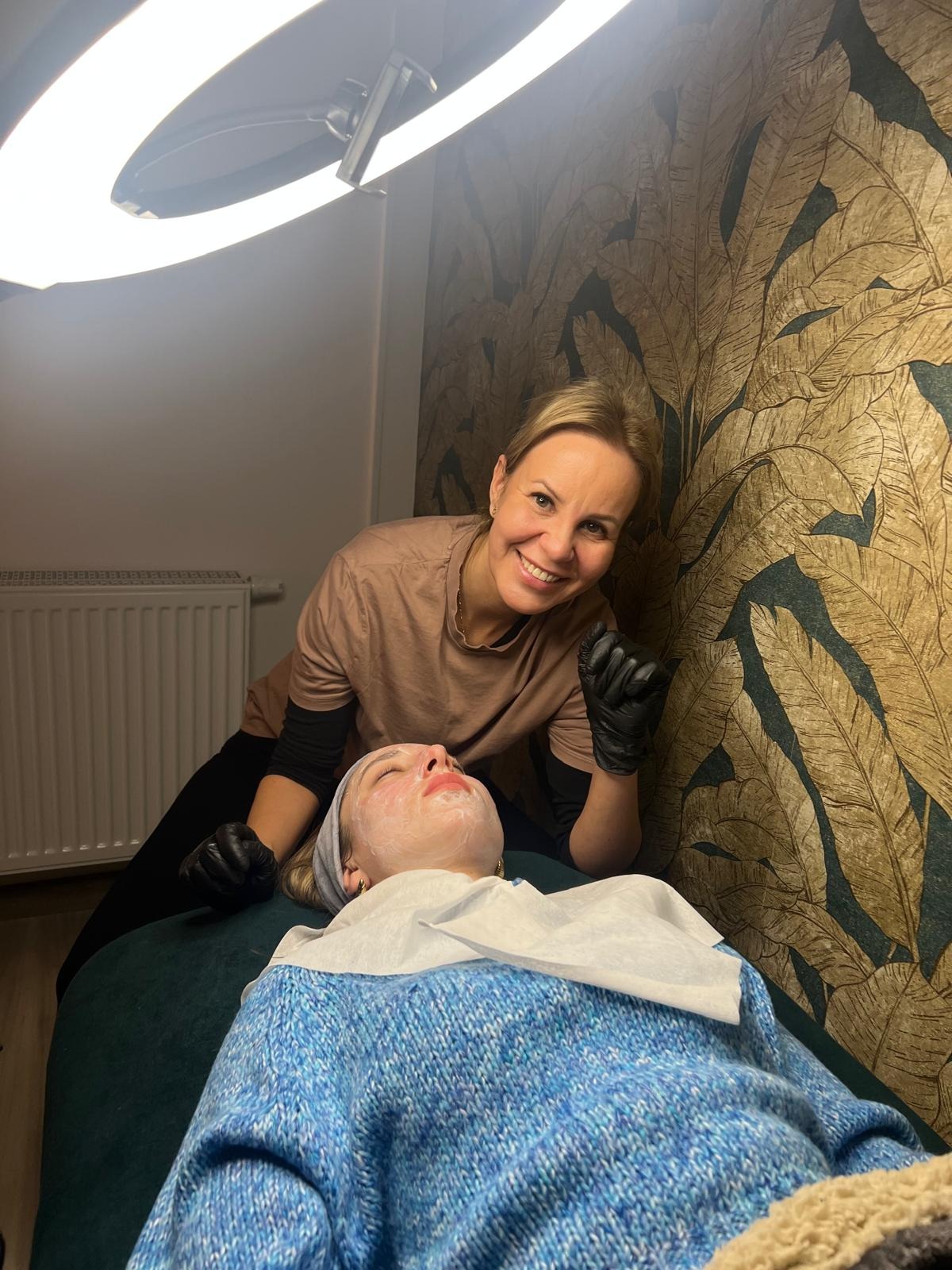 Klientka z nałożoną maską na twarzy leży na zielonym fotelu w trakcie zabiegu upiększającego w Poznaniu, nad nią uśmiechnięta kosmetyczka w czarnych rękawiczkach, w tle ściana z motywem liści...