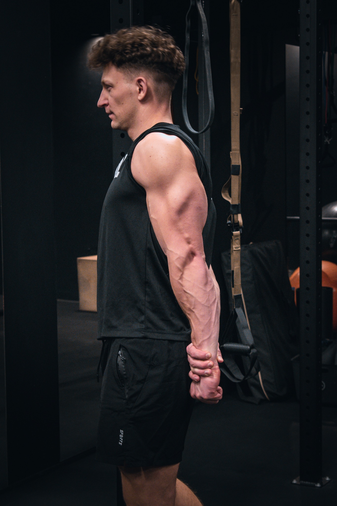 Mężczyzna w czarnym stroju sportowym, profil, widoczne napięte mięśnie ramion i przedramion, dłonie splecione za plecami, siłownia w tle.