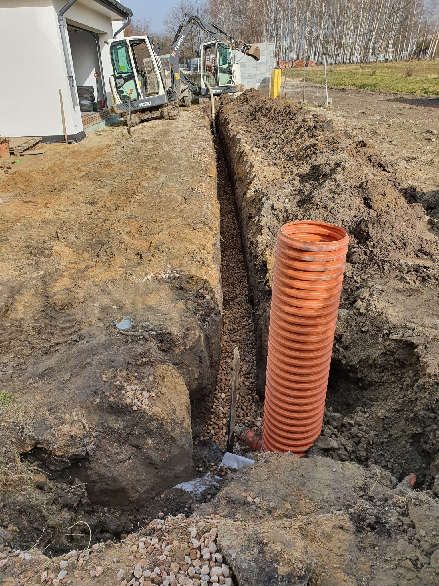 Wykop pod instalację z drenażem wypełniony żwirem, z pomarańczową rurą karbowaną, widoczne dwie koparki Terex TC20 i budynek w tle.