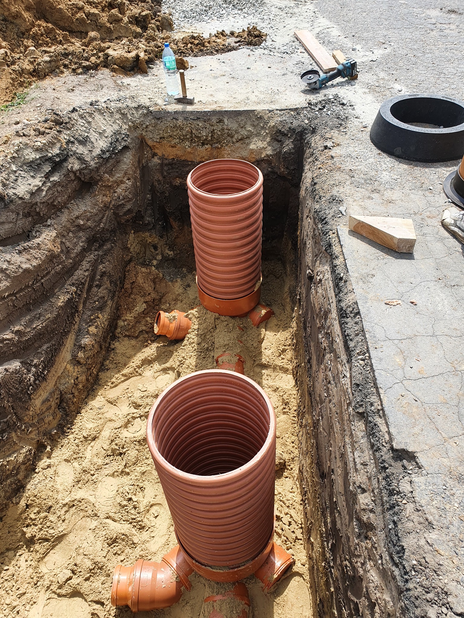 Wykop z piaskowym dnem, w którym znajdują się dwa pionowe odcinki rur karbowanych z tworzywa w kolorze terakoty, połączone kształtkami, na górze wykopu widoczna butelka wody, młotek, szlifierka...