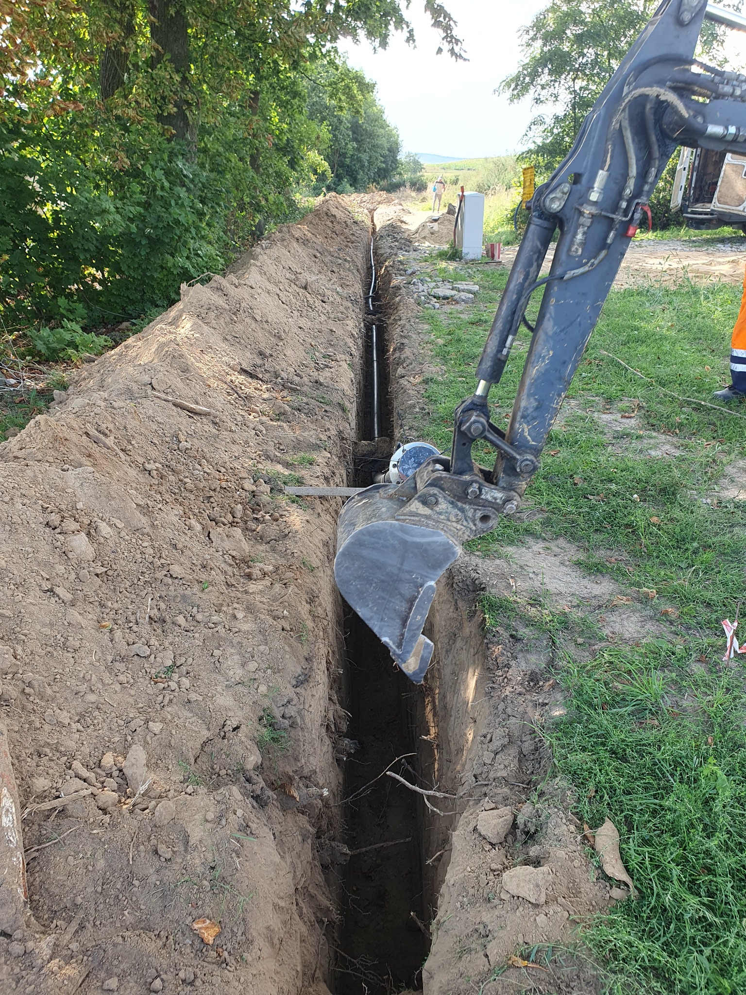 Koparka wykopuje wąski rów w ziemi, widoczna rura w wykopie, w tle zielona roślinność i biała skrzynka.