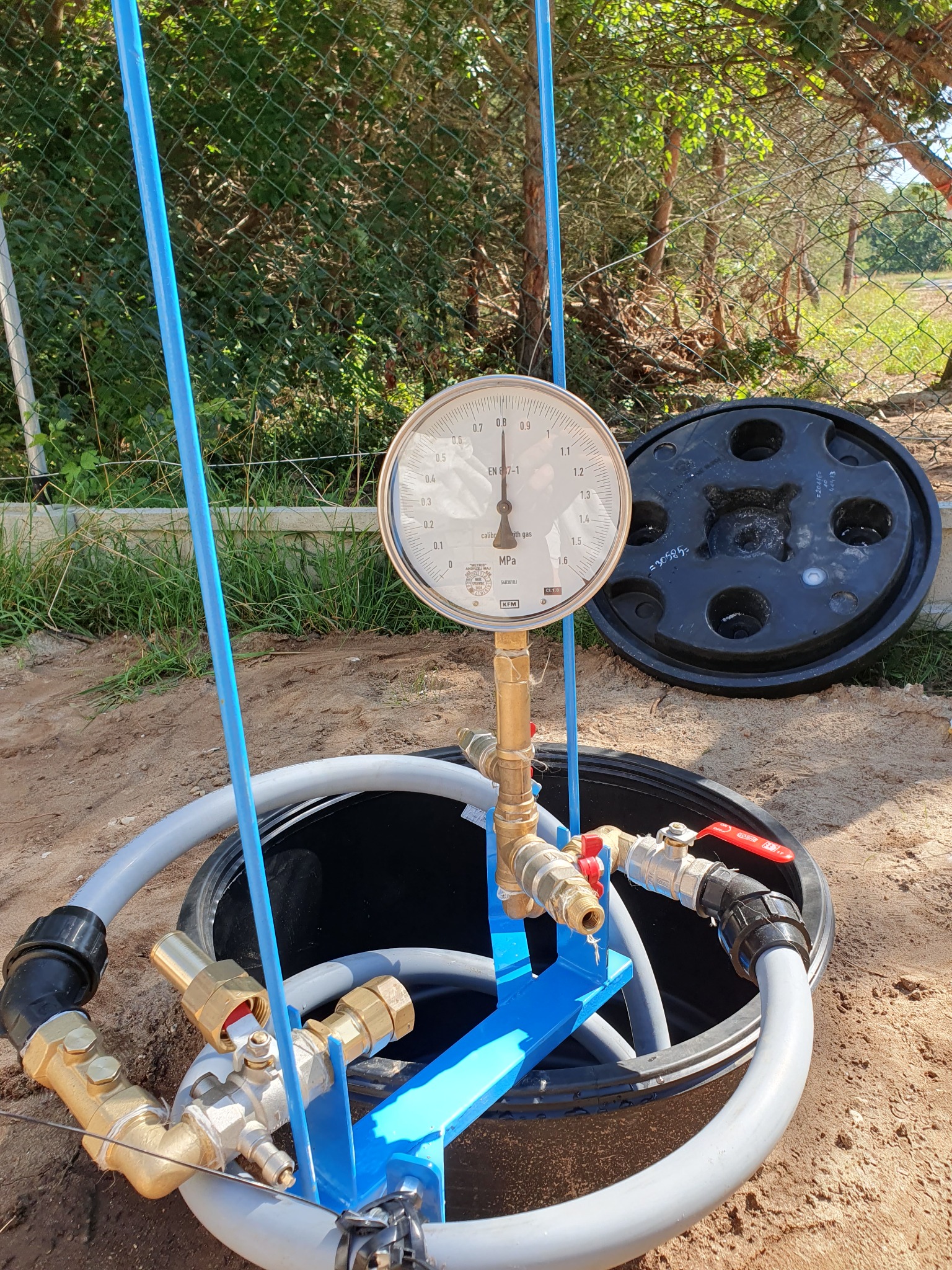 Manometr podłączony do instalacji wewnątrz czarnego zbiornika w ziemi, widoczne rury, zawory i niebieska konstrukcja wsporcza, pokrywa zbiornika obok, w tle trawa i ogrodzenie.