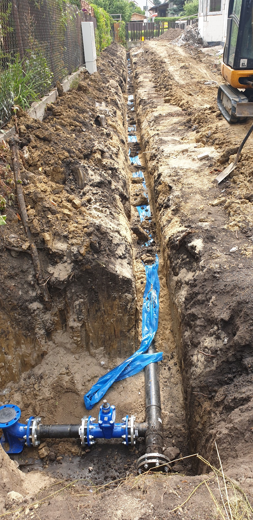 Wykop z ułożonym rurociągiem wodnym zabezpieczonym niebieską folią, widoczne złącza kołnierzowe i zawór odcinający, ziemia wokół wykopu, w tle mini koparka.