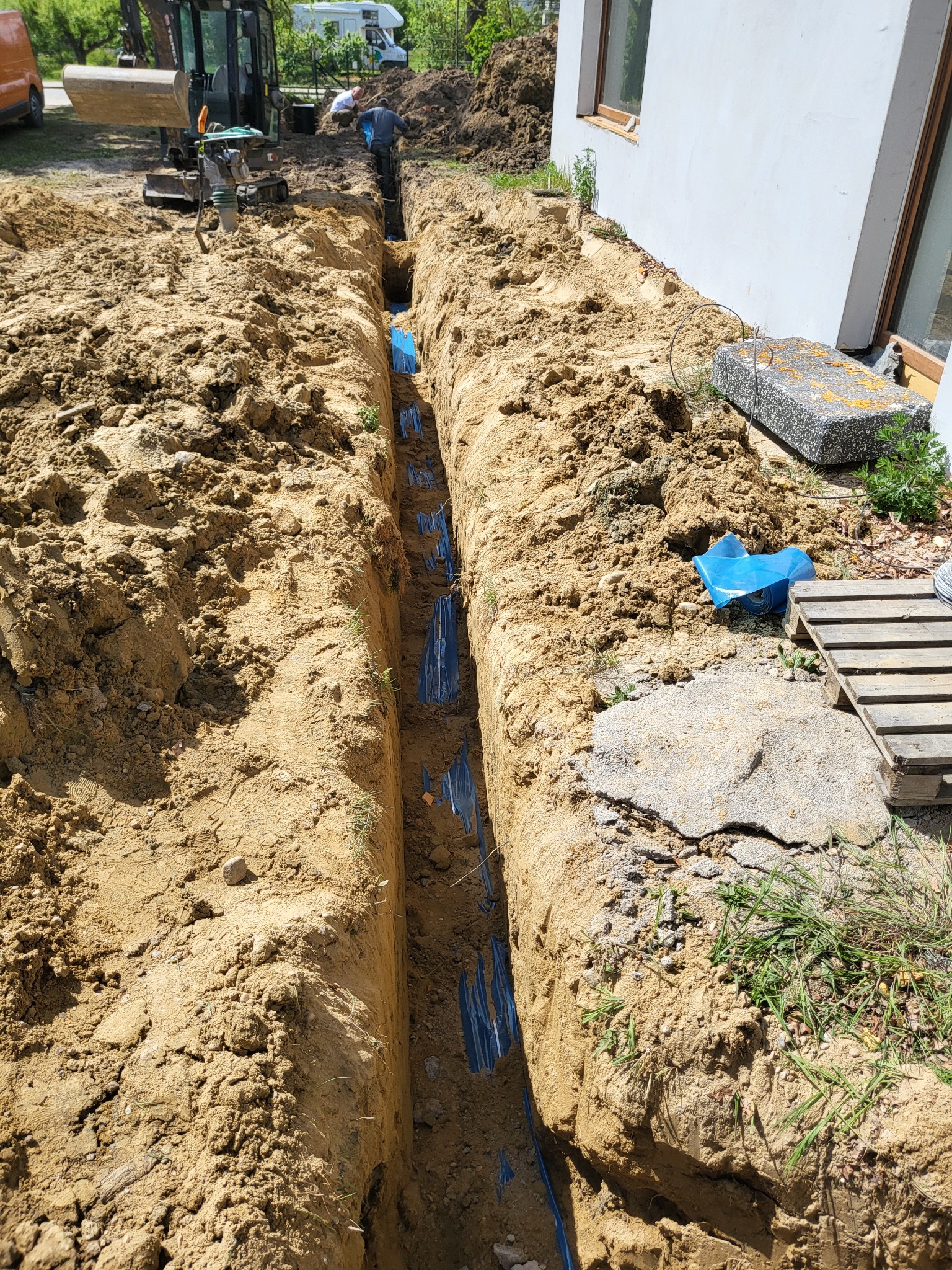 Wykop pod instalację rur w Strzeszowie z widocznym minikoparką i pracownikami, zabezpieczony folią, na tle białego budynku.