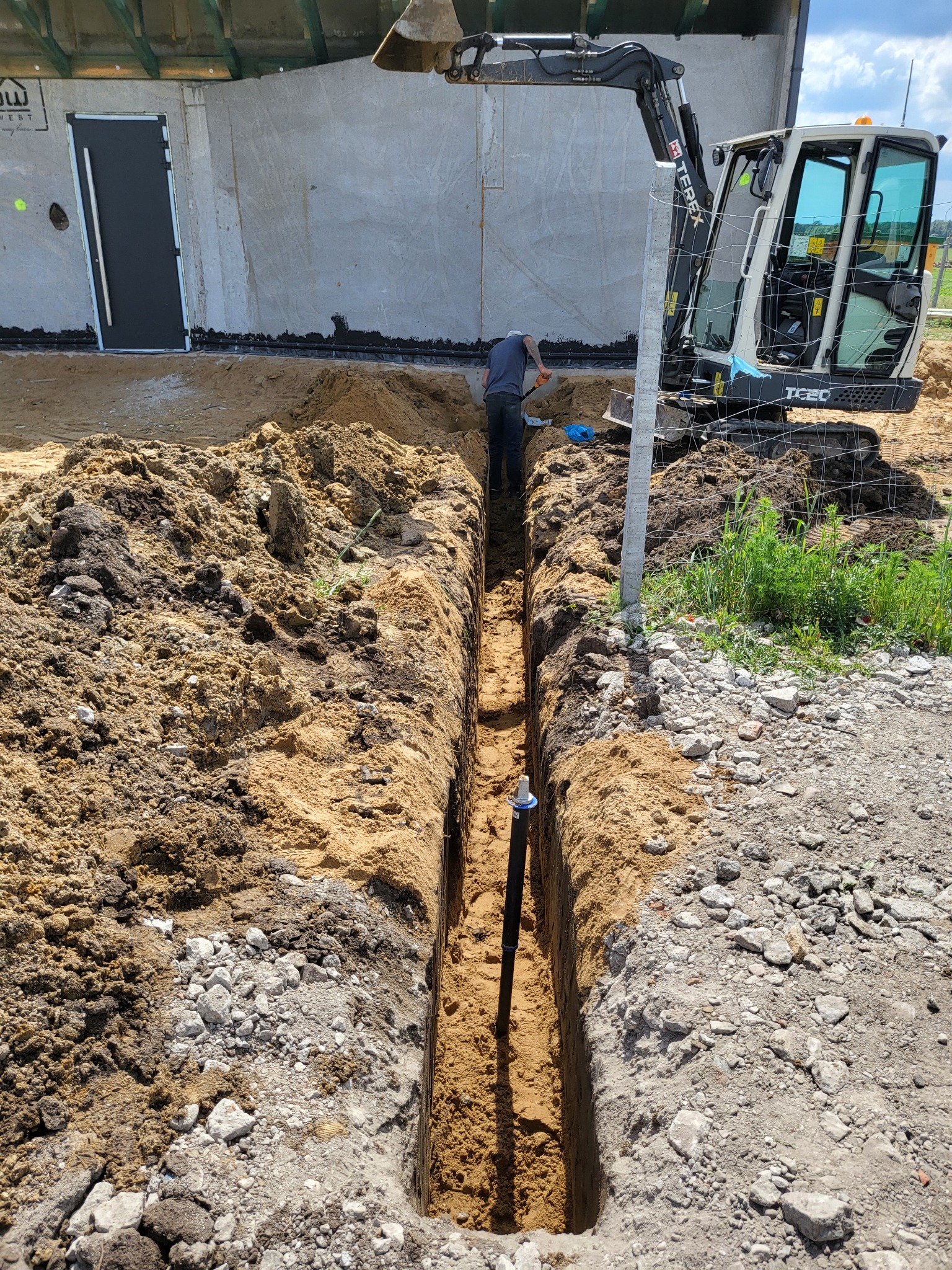 Wykop wąskoprzestrzenny z rurą pionową, wykonywany koparką Terex TC20 i ręcznie przez pracownika w pobliżu budynku w stanie surowym otwartym.