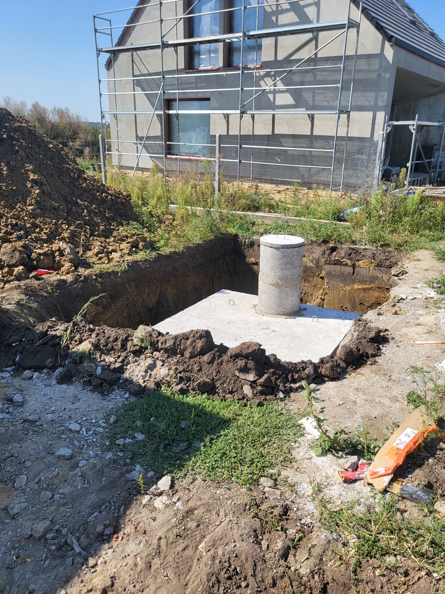 Betonowa konstrukcja z wystającym kominem w wykopie na tle domu w trakcie budowy, widoczne rusztowania.