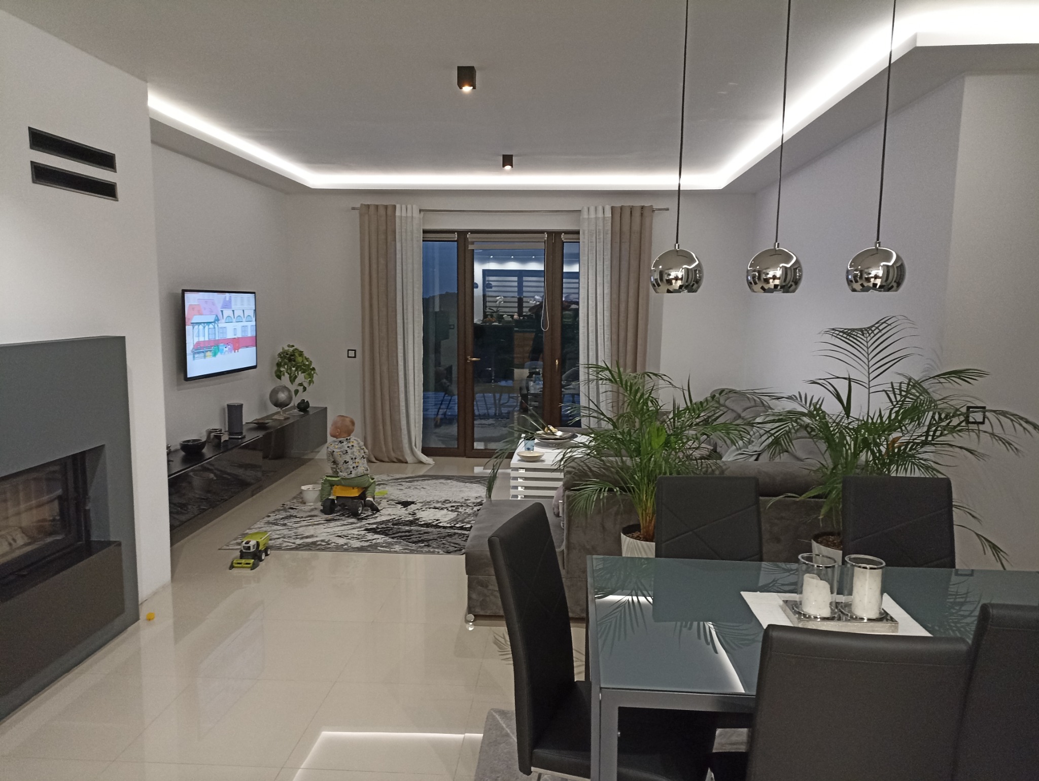 Salon po remoncie: jasne wnętrze z oświetleniem LED, szarym narożnikiem, roślinami i elementami dekoracyjnymi. Widoczny kominek i bawiące się dziecko.