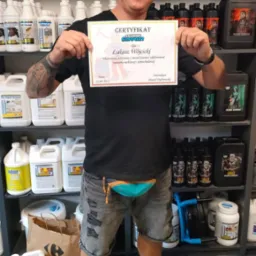 Mężczyzna w czapce trzyma certyfikat ukończenia szkolenia z metod prania i odplamiania tapicerki meblowej i samochodowej, na tle półek z profesjonalnymi środkami czyszczącymi.