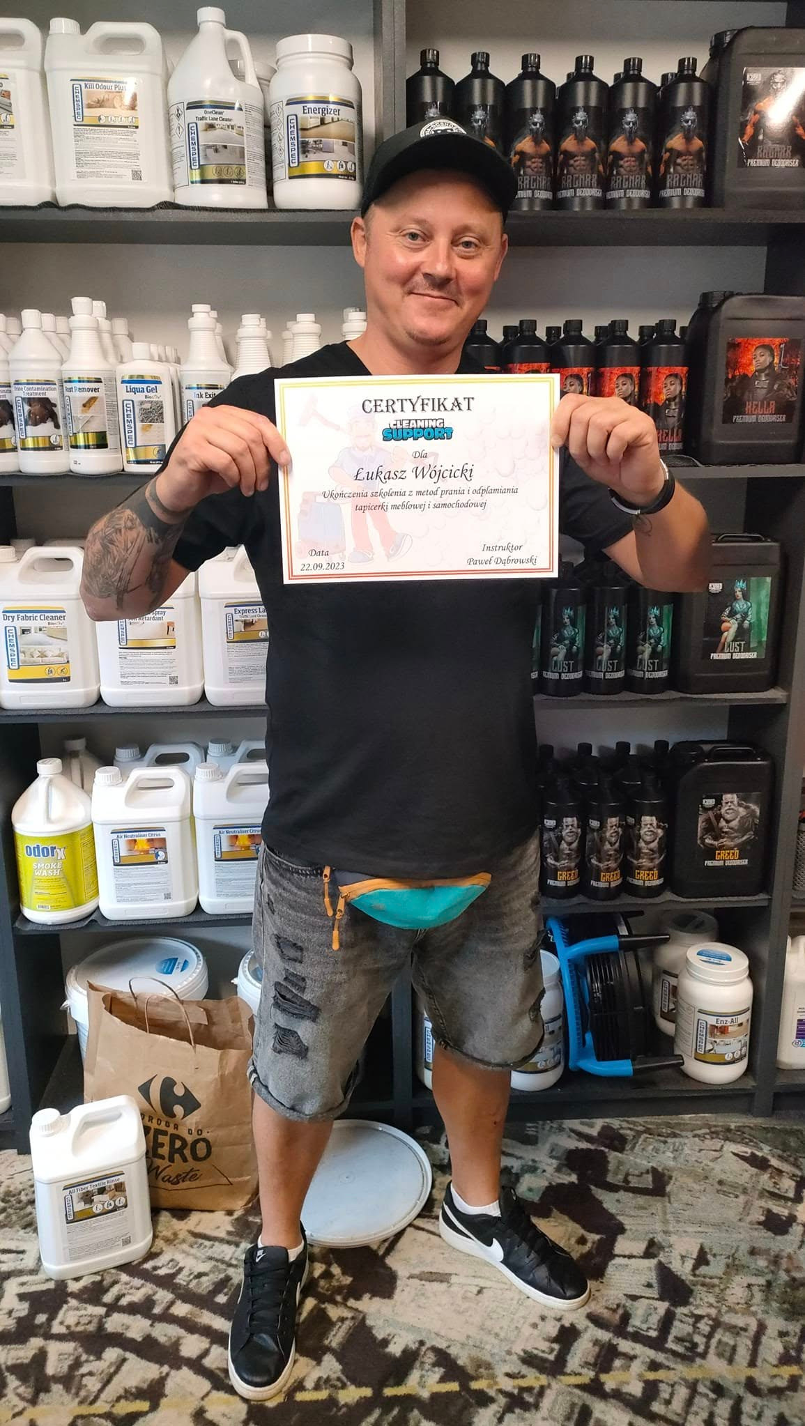 Mężczyzna w czapce trzyma certyfikat ukończenia szkolenia z metod prania i odplamiania tapicerki meblowej i samochodowej, na tle półek z profesjonalnymi środkami czyszczącymi.