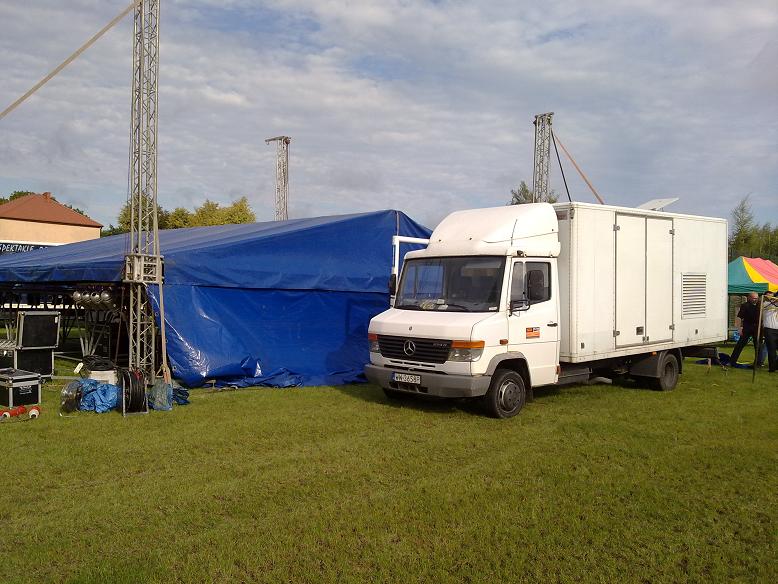 Biały samochód dostawczy z zabudową kontenerową, prawdopodobnie agregatem prądotwórczym, zaparkowany na trawie obok niebieskiego namiotu imprezowego i konstrukcji scenicznej.