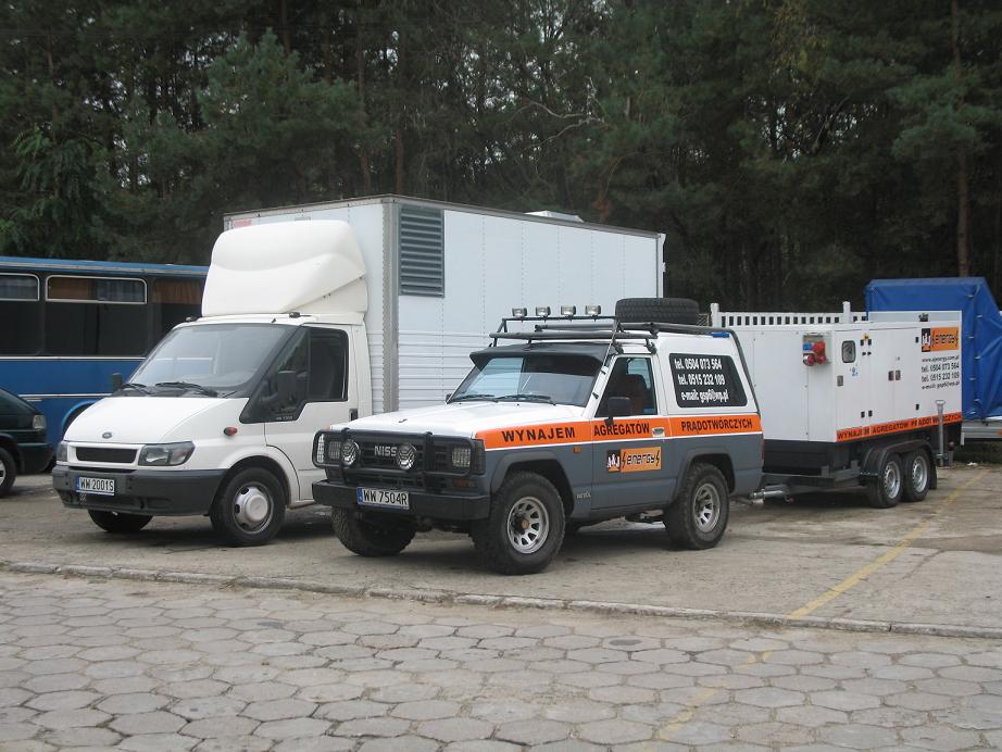 Dwa samochody dostawcze, biały Ford Transit i szaro-biały Nissan Patrol z napisem 'Wynajem Agregatów Prądotwórczych', stojące obok siebie. Do Nissana podłączona jest przyczepa z agregatem...