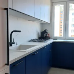 Nowoczesna kuchnia w bieli i granacie: proste szafki, marmurowy blat, czarna armatura i okno z widokiem na miasto. Minimalistyczny design.