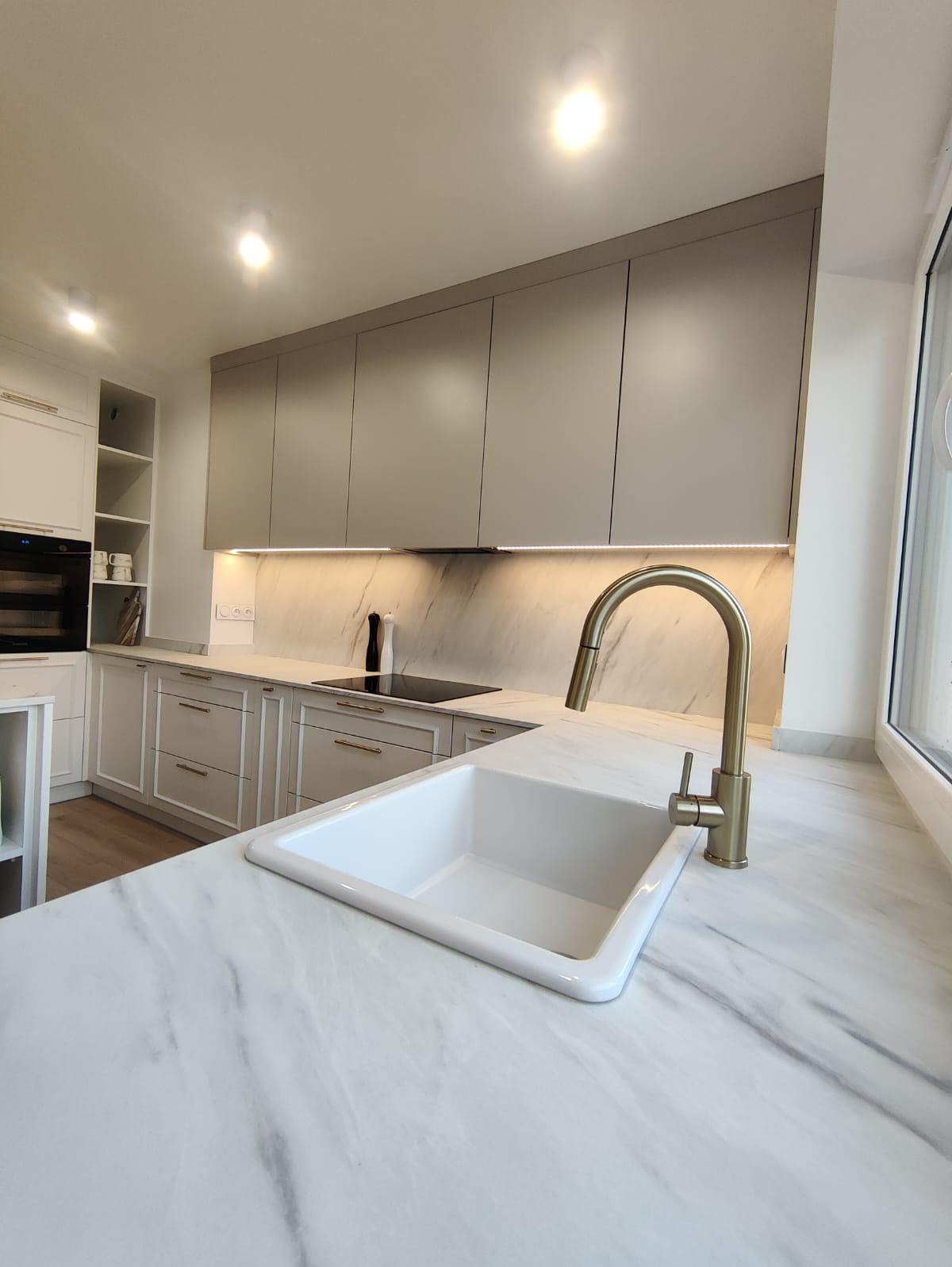 Jasna kuchnia w domu jednorodzinnym. Białe szafki dolne z frezowanymi frontami i złotymi uchwytami, szare szafki górne, marmurowy blat z białym ceramicznym zlewem i złotą baterią. Oświetlenie LED...