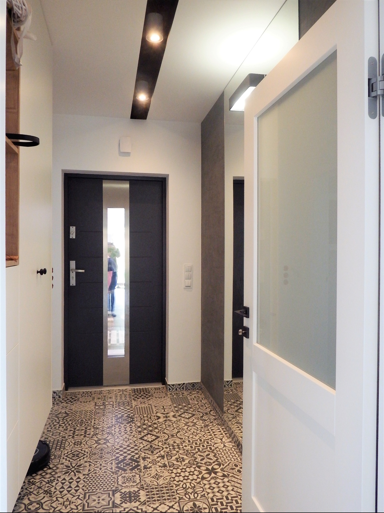 Nowoczesny przedpokój z czarnymi drzwiami wejściowymi z przeszkleniem, wzorzystą podłogą w stylu retro i białymi drzwiami wewnętrznymi z matowego szkła.