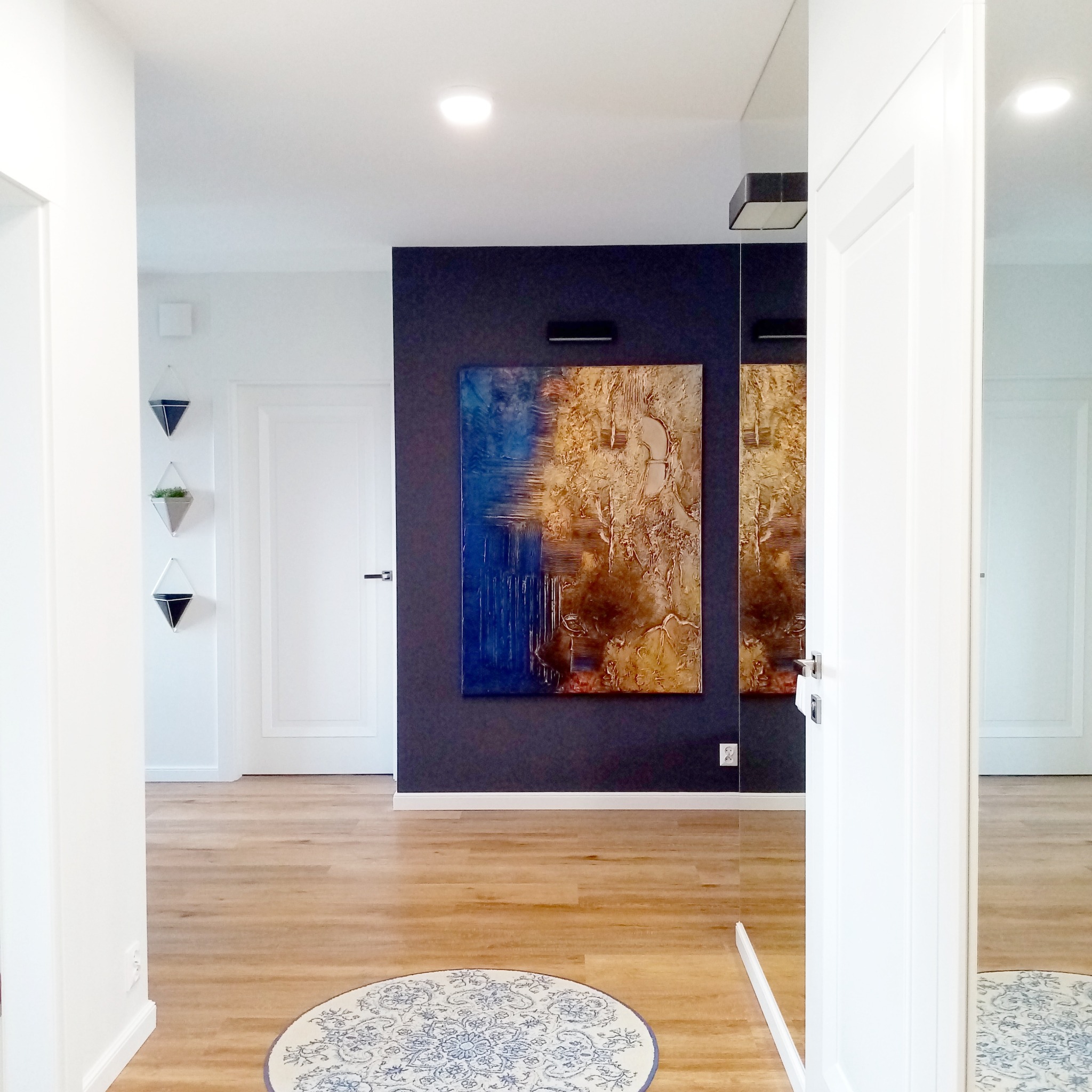 Nowoczesny przedpokój z podłogą z paneli, okrągłym dywanem w kwiaty i abstrakcyjnym obrazem na ciemnej ścianie. Minimalistyczne doniczki wiszące.