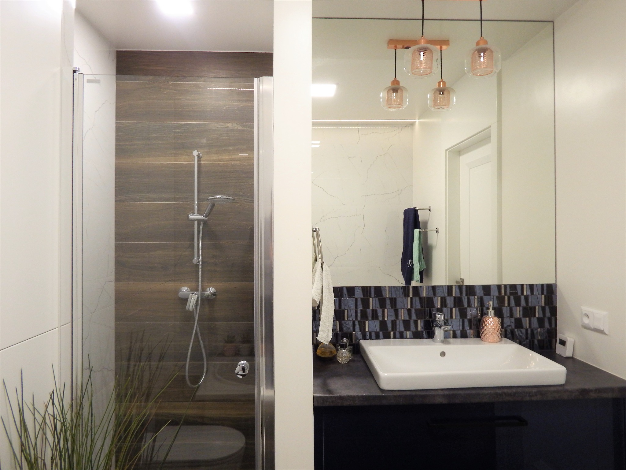 Nowoczesna łazienka z kabiną prysznicową i umywalką, lustro odbija wnętrze. Drewnopodobne płytki i szklane lampy dodają elegancji.