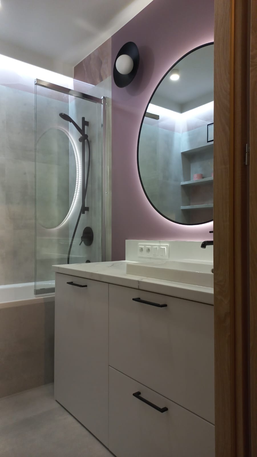 Nowoczesna łazienka: przeszklona kabina prysznicowa, szafka z umywalką, okrągłe lustro z podświetleniem LED i designerska lampa na fioletowej ścianie.