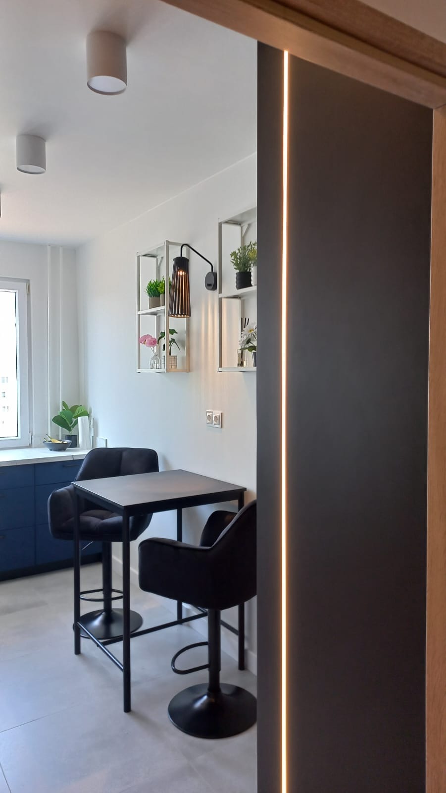 Nowoczesna aranżacja małej jadalni z czarnym stolikiem barowym i krzesłami. Na ścianie półki z dekoracjami, oświetlenie LED. Minimalistyczny design.