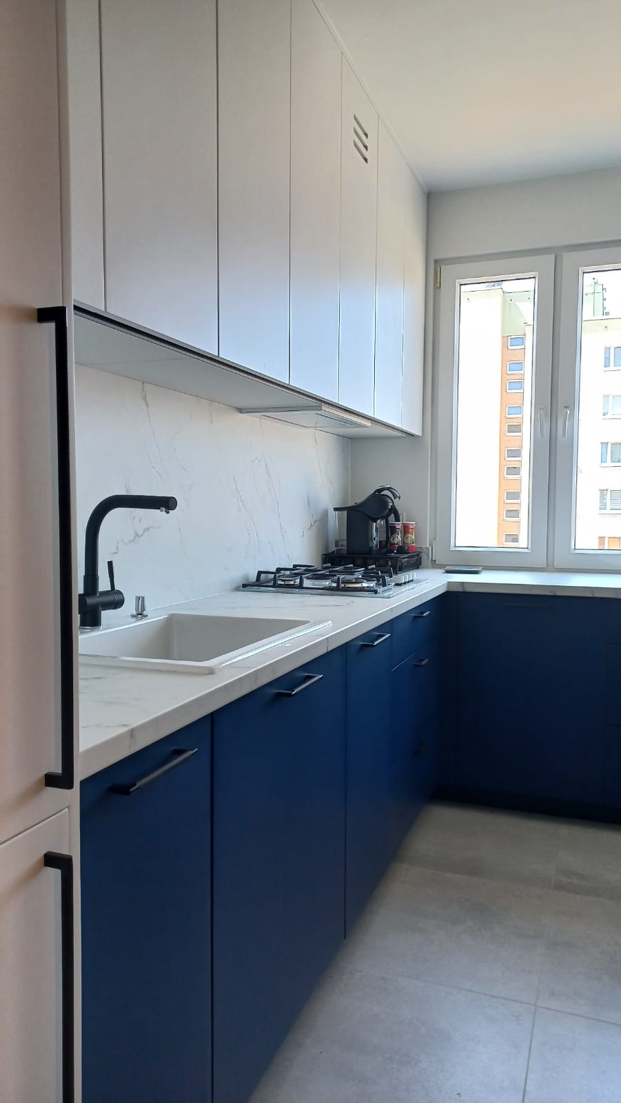 Nowoczesna kuchnia w bieli i granacie: proste szafki, marmurowy blat, czarna armatura i okno z widokiem na miasto. Minimalistyczny design.