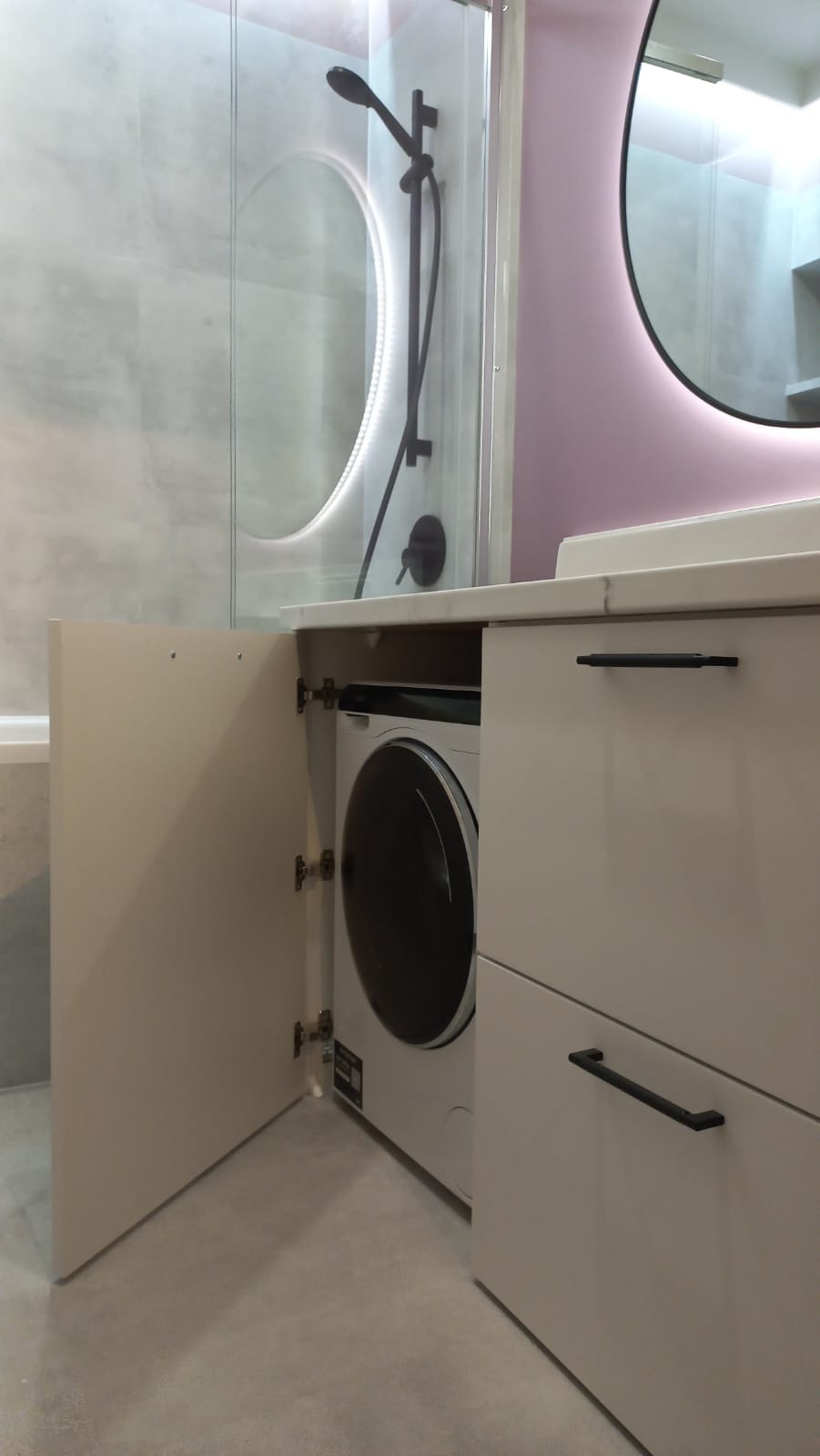 Nowoczesna łazienka z pralką ukrytą w szafce pod umywalką, okrągłe lustro z podświetleniem LED, kabina prysznicowa z czarną armaturą.