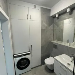 Nowoczesna łazienka w Elblągu z szarymi płytkami imitującymi marmur, białą szafką zabudowaną nad pralką automatyczną oraz wiszącą toaletą i umywalką z szafką.