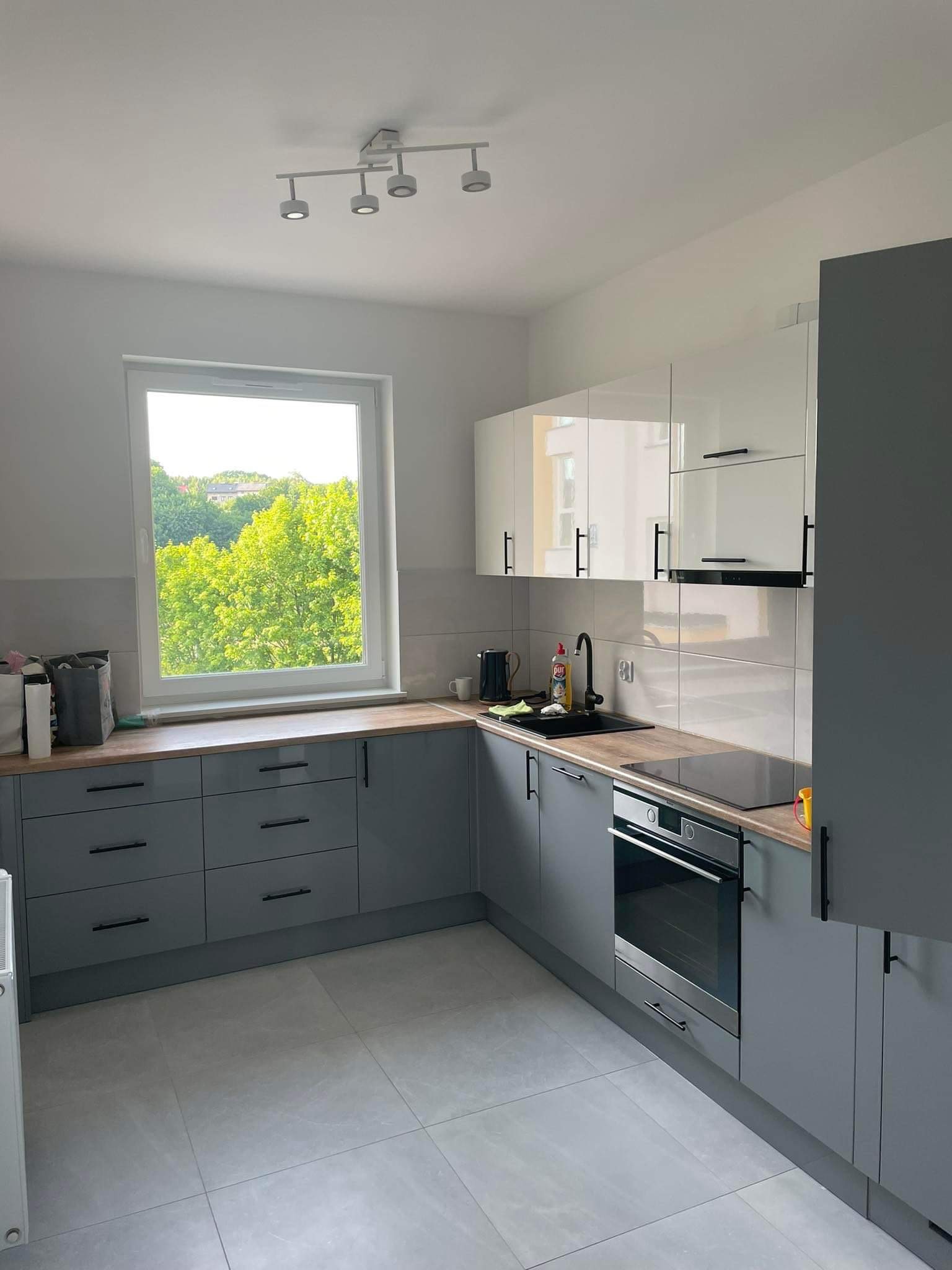 Nowoczesna kuchnia w odcieniach szarości i bieli z drewnianym blatem, czarną armaturą i widokiem na zieleń za oknem.