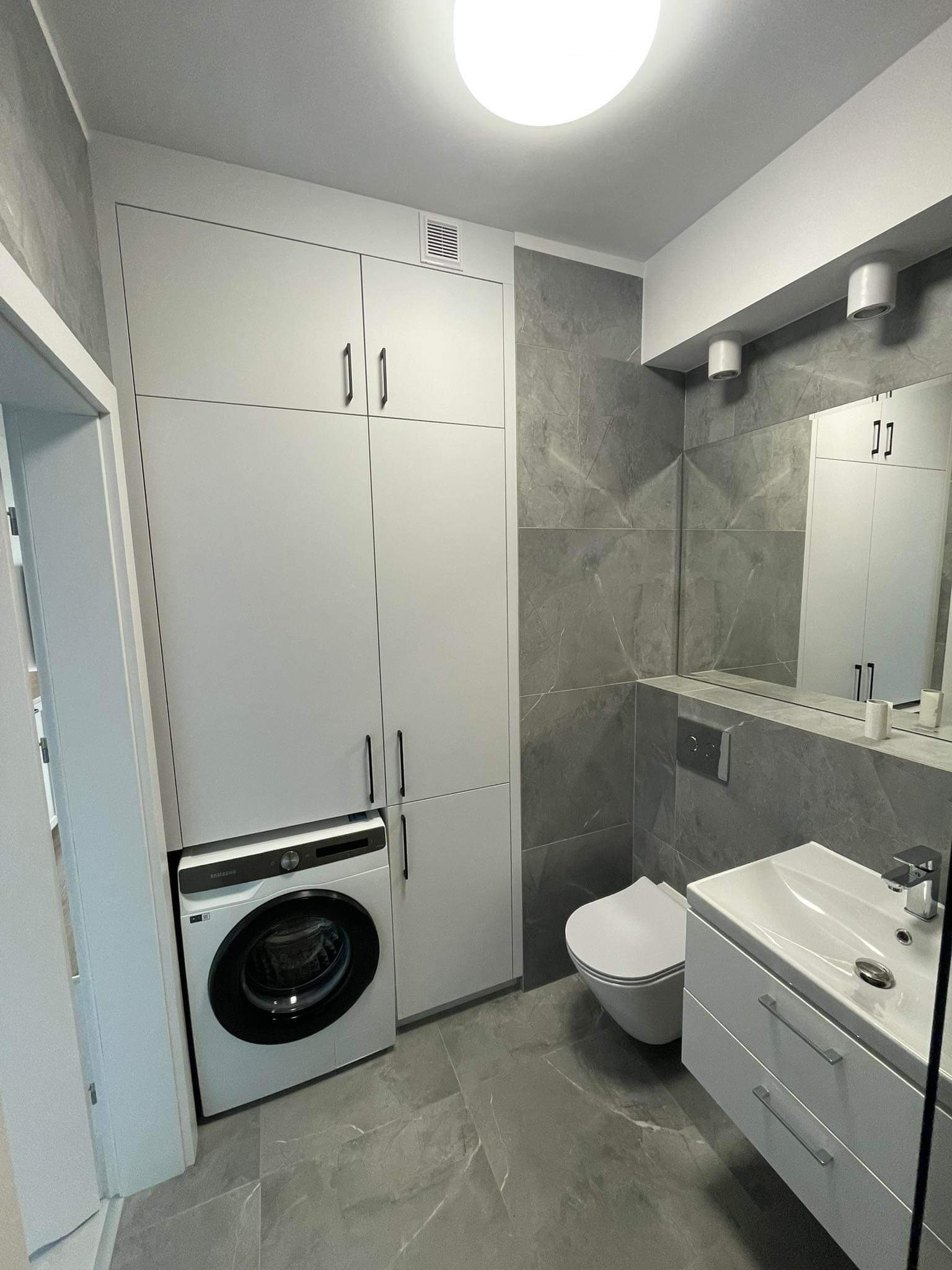 Nowoczesna łazienka w Elblągu z szarymi płytkami imitującymi marmur, białą szafką zabudowaną nad pralką automatyczną oraz wiszącą toaletą i umywalką z szafką.