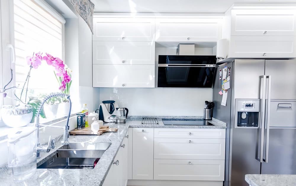 Biała kuchnia na wymiar z granitowym blatem, nowoczesnym okapem i stalową lodówką. Na blacie kwiaty i akcesoria kuchenne. Jasne wnętrze z oknem.