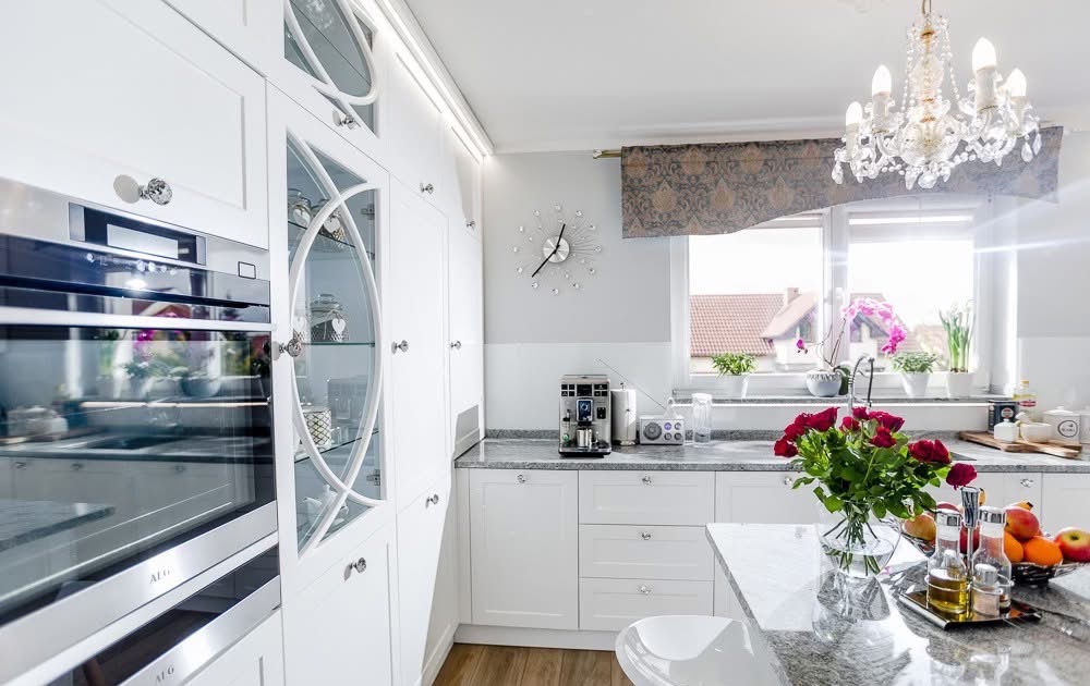 Jasna, nowoczesna kuchnia z białymi szafkami, przeszklonymi witrynami, blatem z szarego kamienia i bukietem czerwonych róż na wyspie.