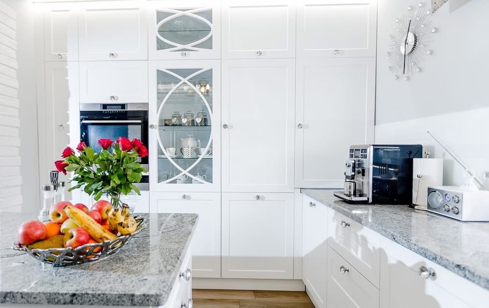 Nowoczesna, biała kuchnia z granitowym blatem i dekoracyjnymi szklanymi frontami szafek. Na blacie misa z owocami i bukiet czerwonych róż.