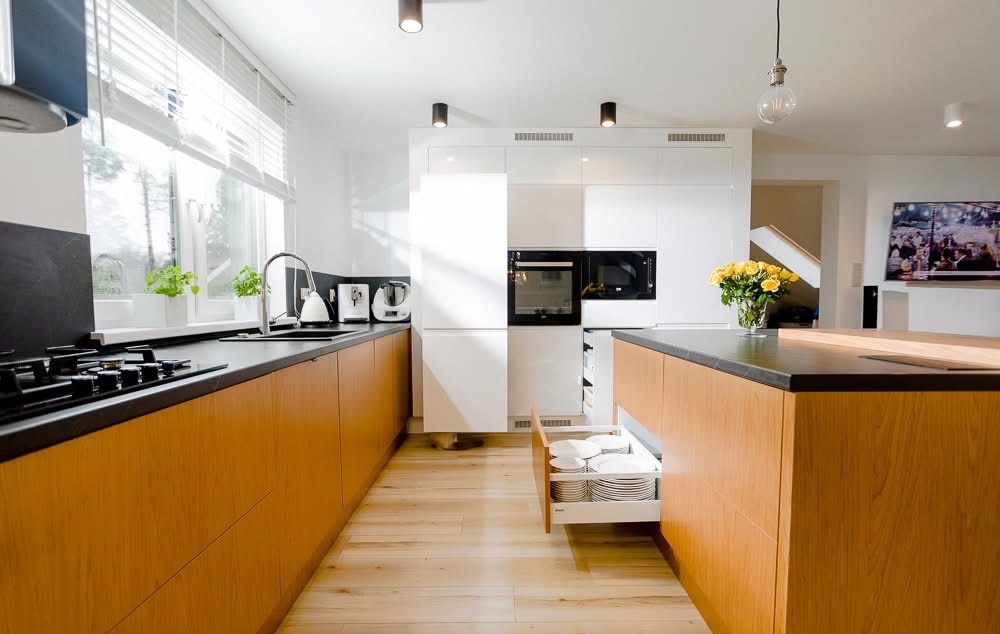 Jasna kuchnia z drewnianymi frontami szafek, czarnym blatem i otwartą szufladą z białymi talerzami. Nowoczesne oświetlenie i minimalistyczny design.