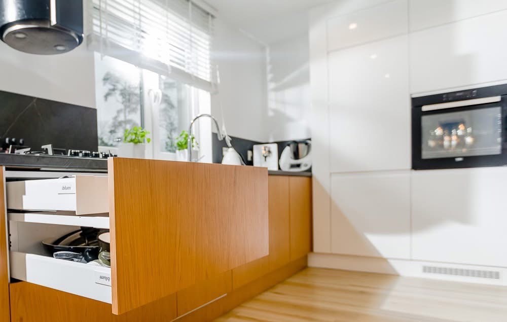 Nowoczesna kuchnia z drewnianymi frontami i otwartymi szufladami Blum, jasne wnętrze z oknem i sprzętami AGD. Detale wykończenia i oświetlenie.